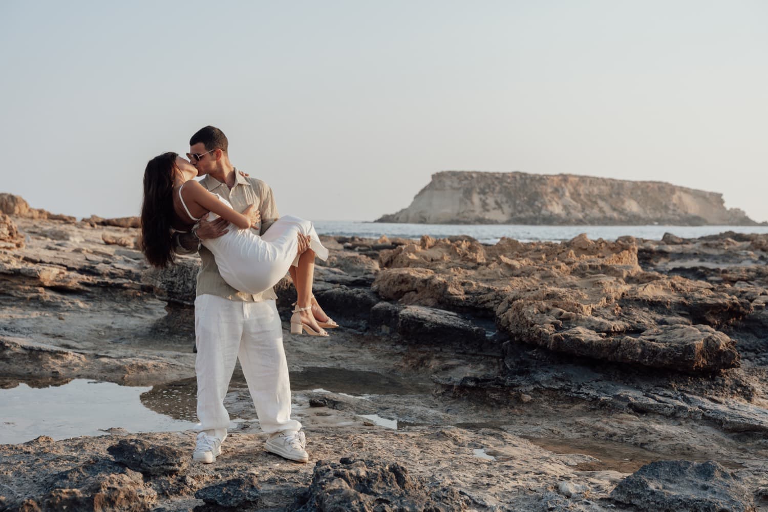 A couple embracing on the rocks above the sea, with an islet beyond, on the Cyprus coast
