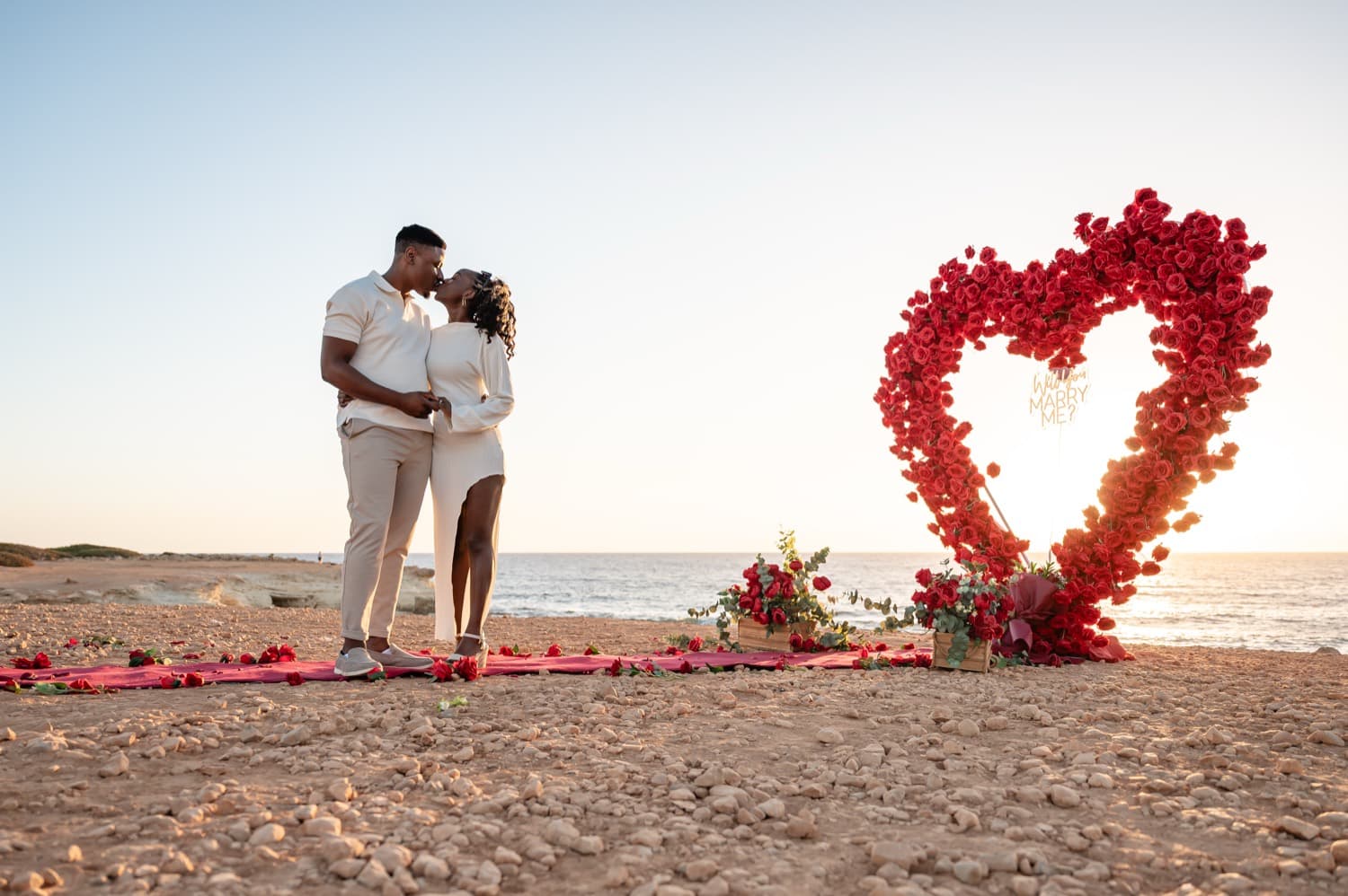 A couple kissing before a red rose heart arch, backlit by the setting sun over the sea in Cyprus